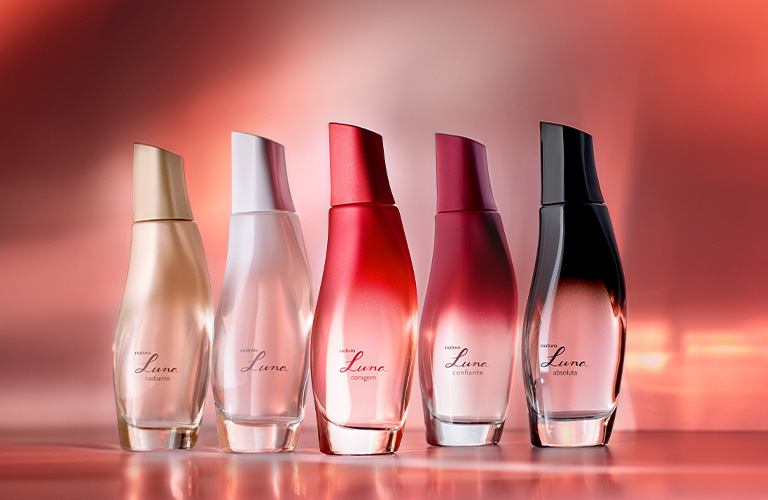 Fotografía de frascos de perfume alineados sobre una superficie reflectante. El fondo muestra tonos degradados en rojo y rosa.